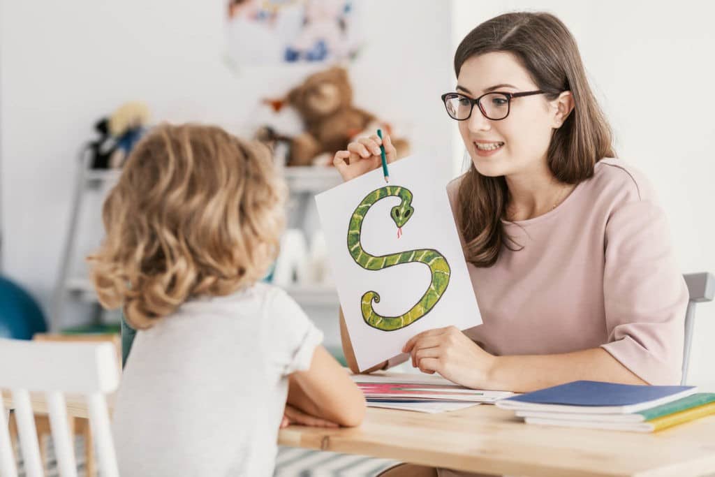 A teacher sitting with a young student holding up a drawing of a snake in the shape of the letter ‘S’.