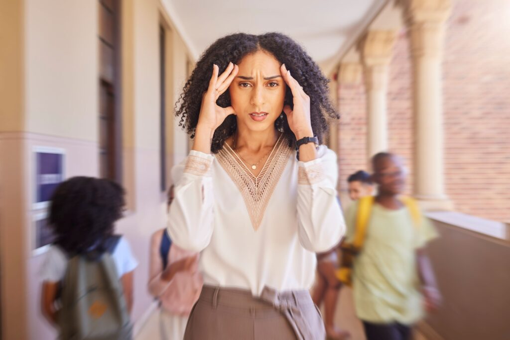 A teacher stands in the hallway with her hands on her head, looking stressed as students zoom around her.