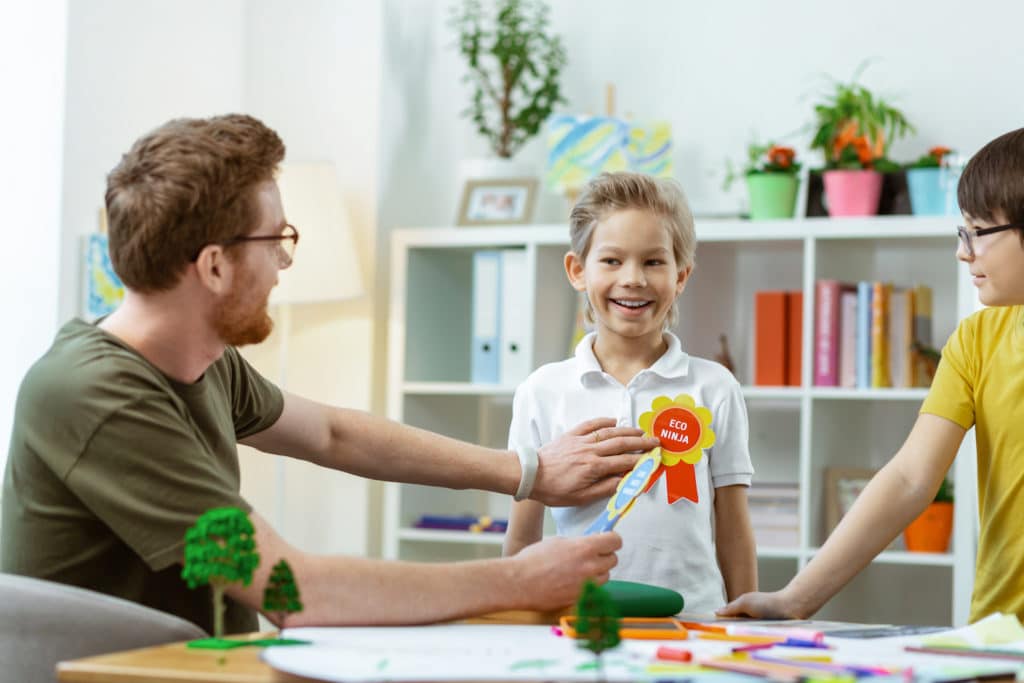 Smiling boy receiving a ribbon from a teacher in a classroom.