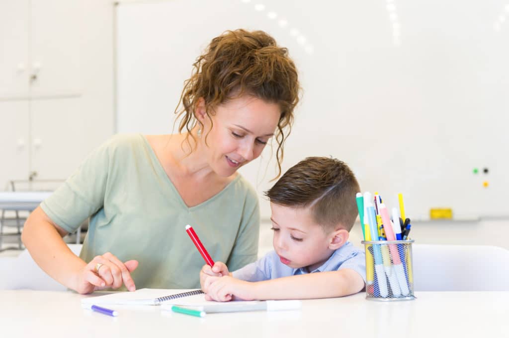 Teacher sitting with a young child coloring in a notebook.