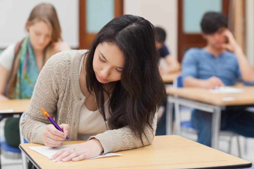 Girl sitting at a desk writing a piece of paper in a classroom.