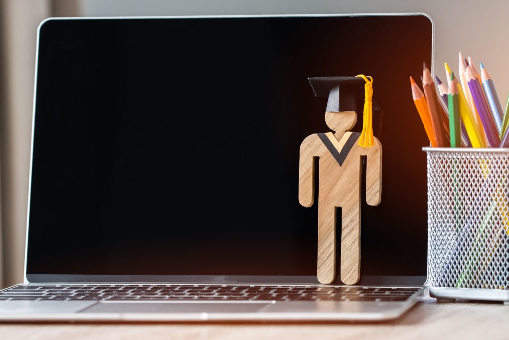 A wooden cut-out of a person stands on a laptop wearing a graduation cap.