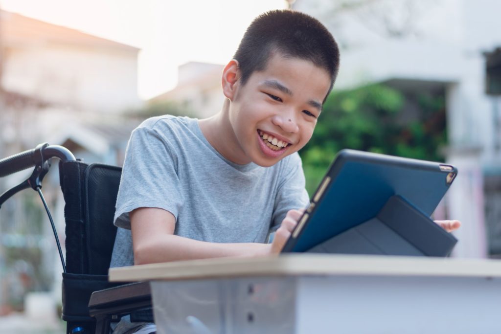 A special education students sits in his wheelchair, smiling at his iPad.