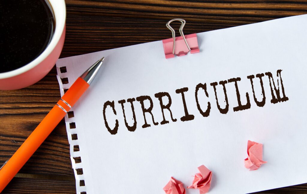 The word “curriculum” is written on a piece of paper, sitting next to a coffee cup, representing curriculum writing.