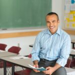 A teacher sits on a desk in his classroom, smiling and holding a stack of papers.