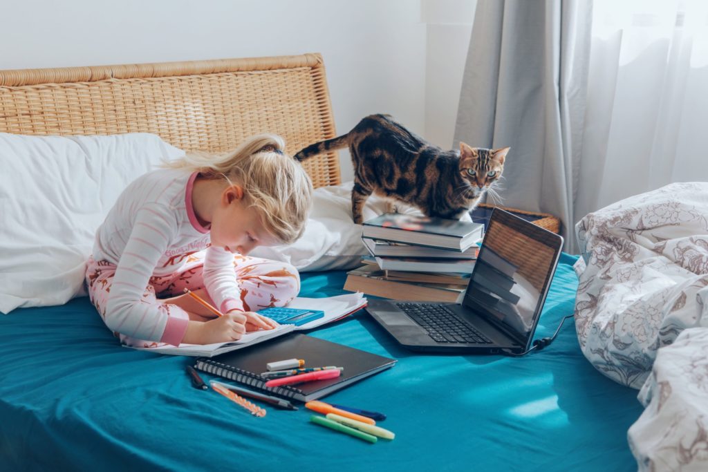 A young student sits in her bed with her books and laptop, doing remote learning.