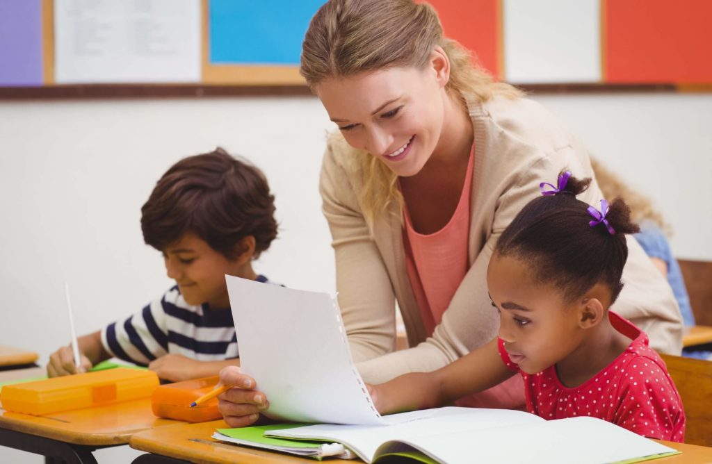 A female teachers looks over a student’s paper in an elementary classroom.