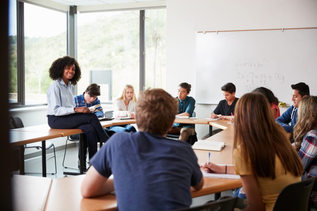 A female high school teacher sits with a class of students, in the middle of a lesson.