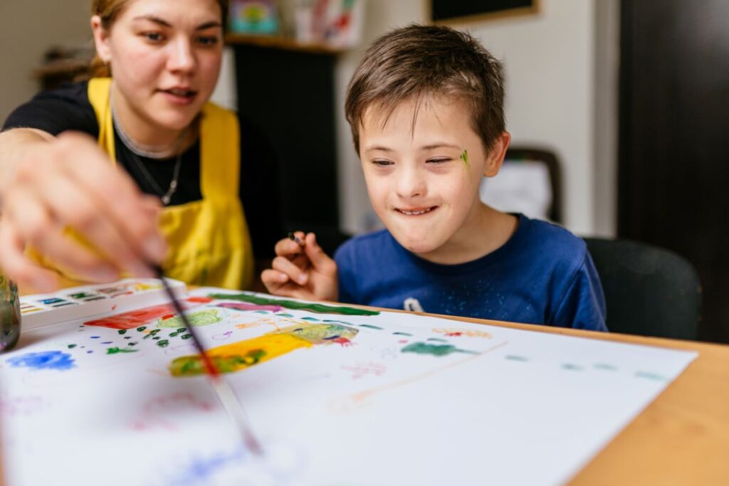 A young teacher sits with a special education student as he paints.