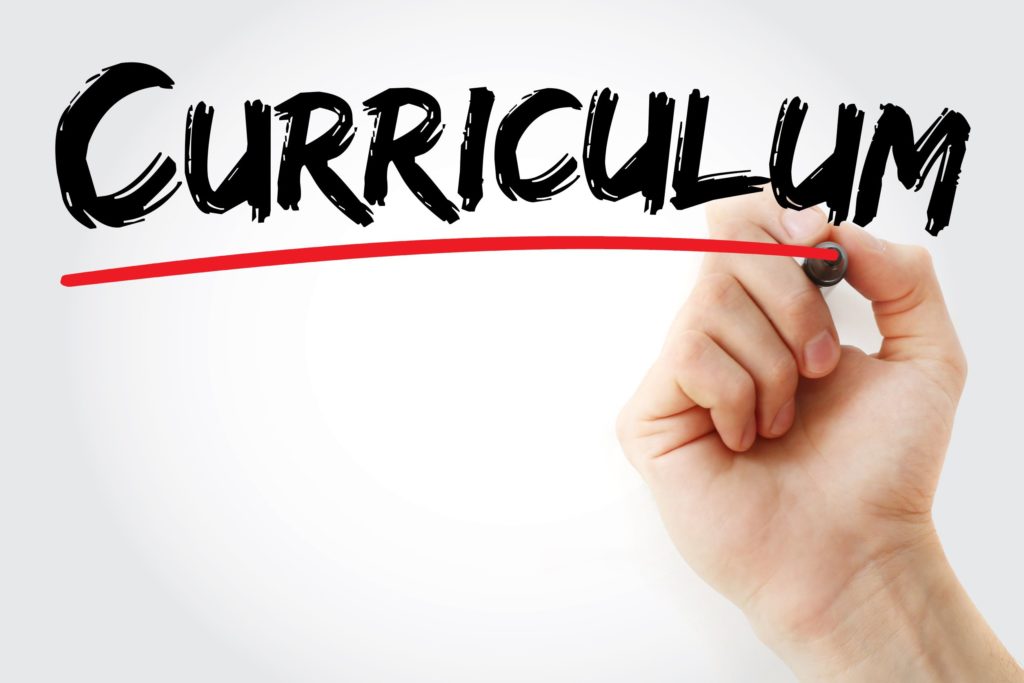 A hand writes the word “curriculum” and underlines it with a marker.
