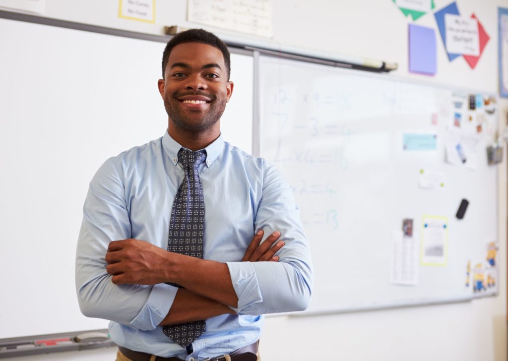 Male teacher smiling and standing in the front of a classroom with his arms crossed.