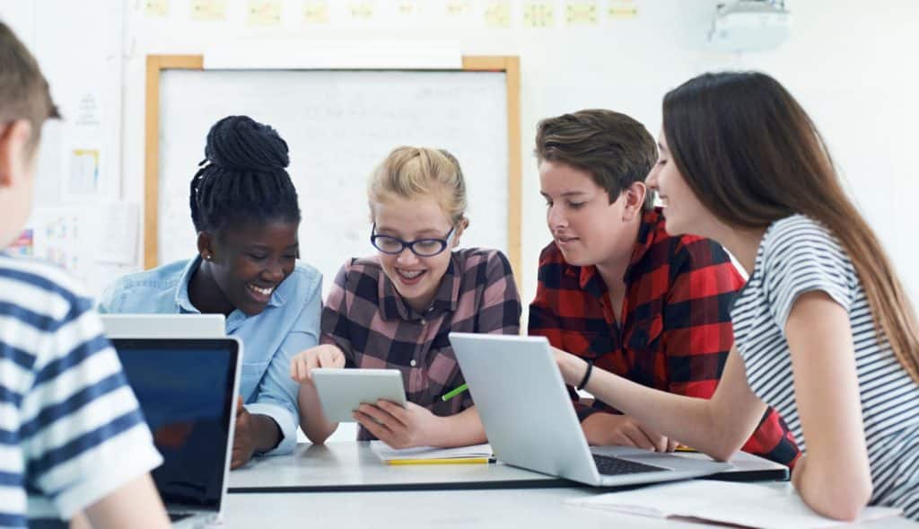 Diverse group of students sitting at a table working together with a tablet and laptops.