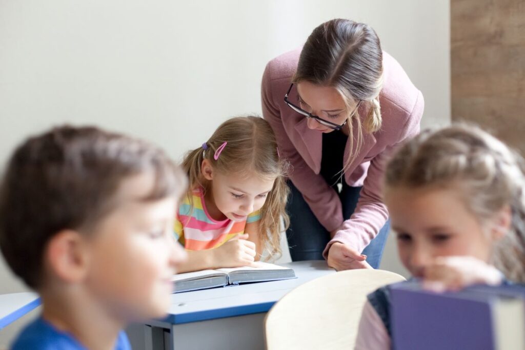 A literacy consultant bends down and helps a student with her reading.