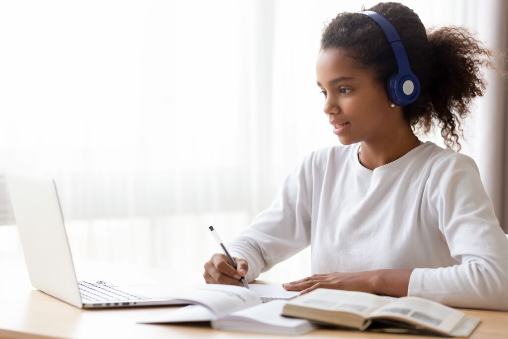 A young girl wears headphones and take notes in a notebook, looking at her laptop.