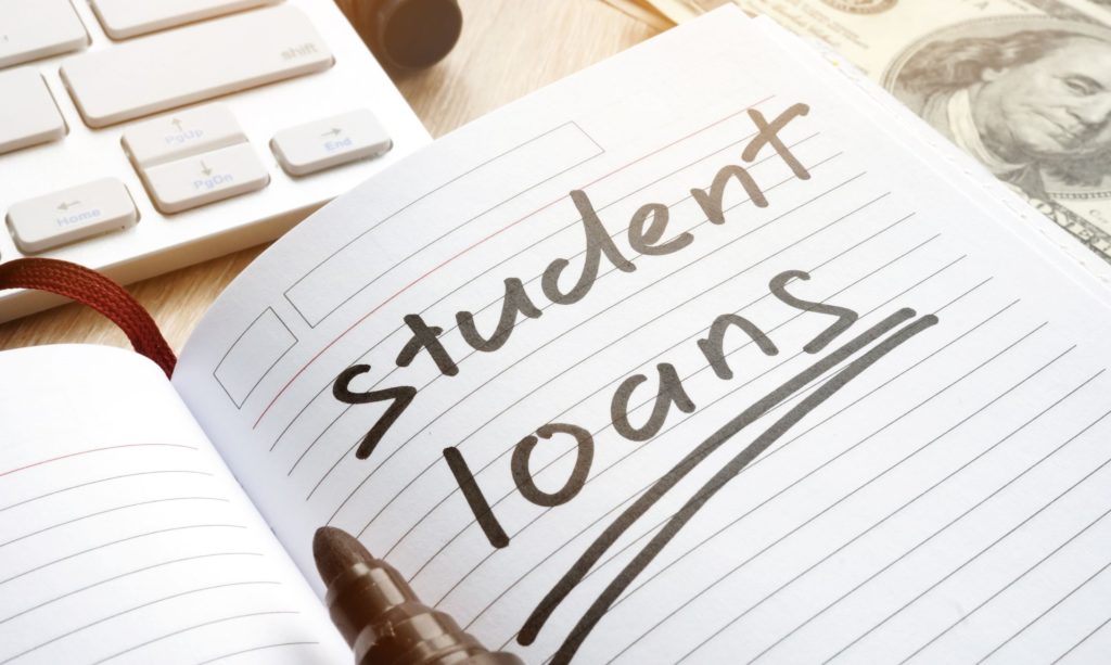 Student Loans’ written in a notebook next to a keyboard and dollar bills.