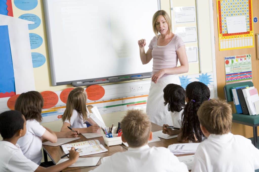 Teacher in front of smart board addressing kids at a table.