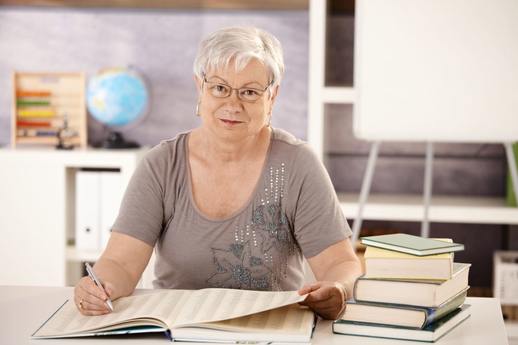 Older teacher sitting at a desk in a classroom searching for something in a book.