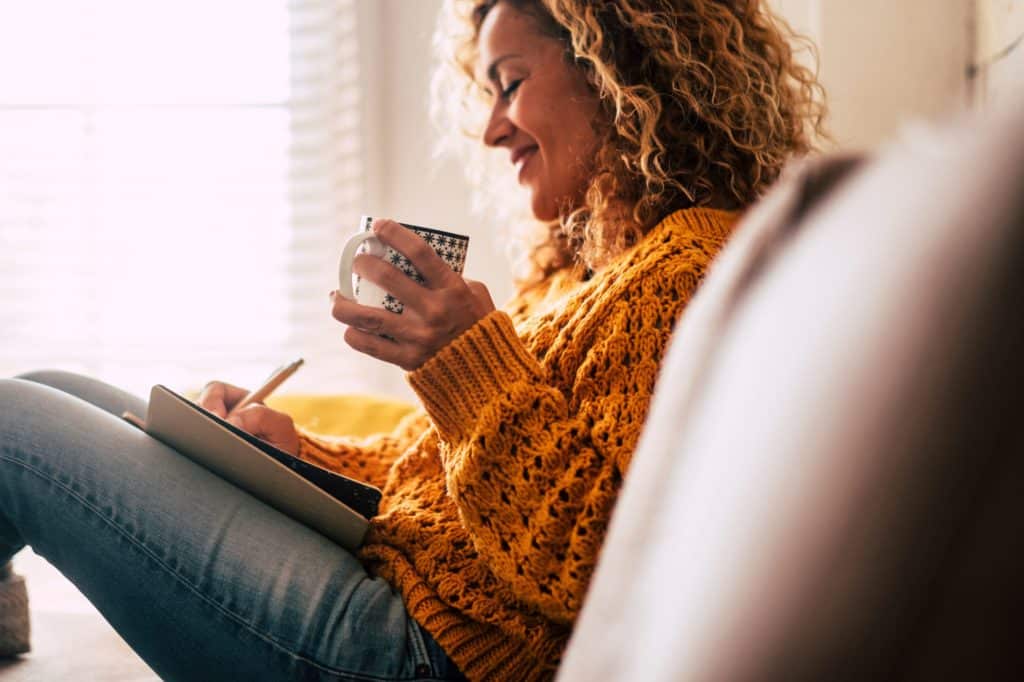 Smiling woman sitting on a couch with a cup of tea writing in a notebook.
