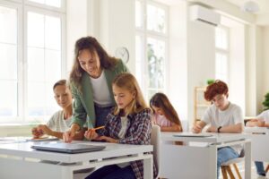 A smiling teachers helps her students with an assignment they are doing at their desks.