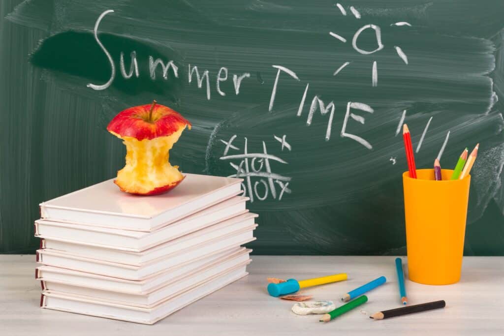 Portrait of a chalkboard that reads “summertime” written in chalk behind an apple core sitting on a stack of books.