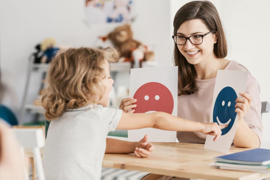 Woman sitting at a table with a young child holding up papers with emoticons.