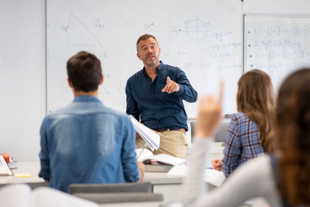 A math teacher points to a student who is raising their hand to answer a question.