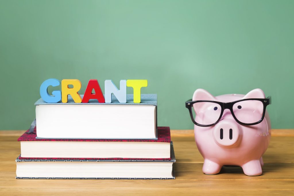 A piggy bank sit in from of a chalkboard; colorful letters read “grant” on top of a stack of books.