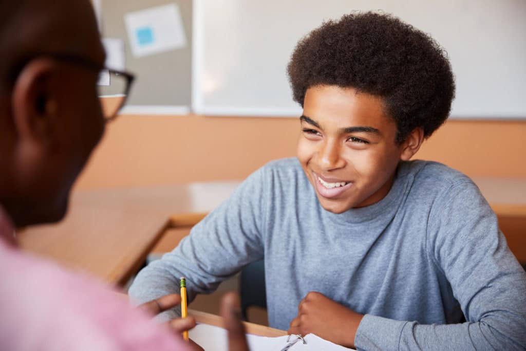 Young boy smiling and sitting at a desk across from a teacher.