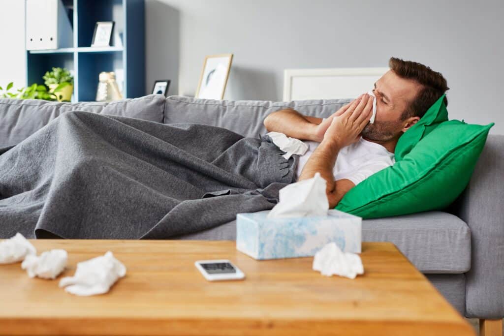 A sick man lays on his couch, blowing his nose and taking a sick day from work.