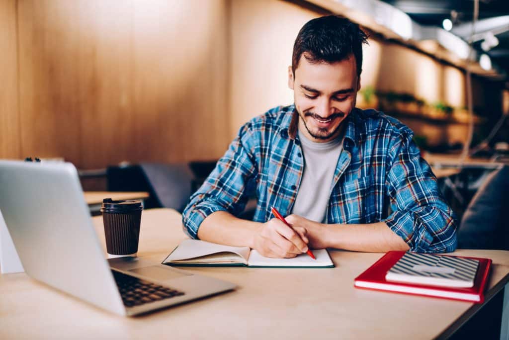 A smiling man works on a project, writing down information in a notebook.
