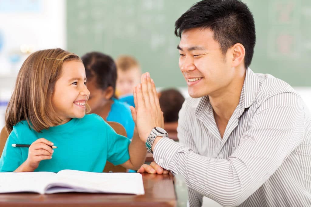 A smiling student and smiling teacher high-fiving at a desk.