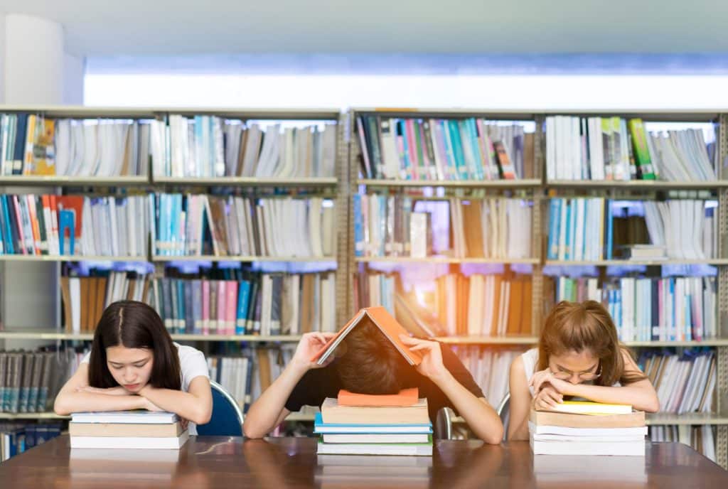 Multiethnic students sit in a library, laying their heads down on their school books in a stressed manner.