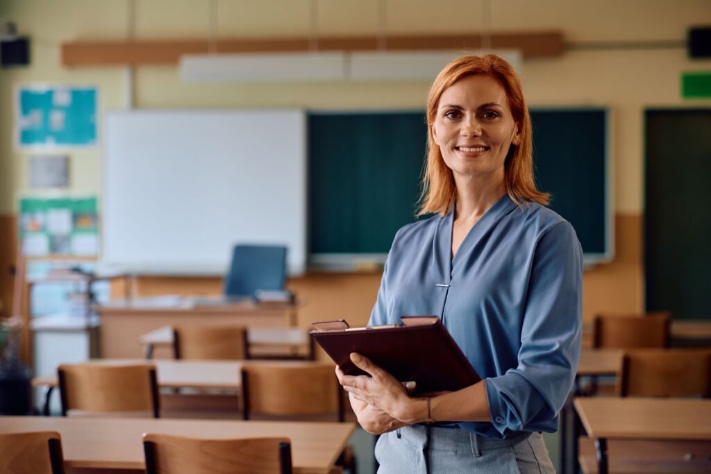 A portrait of a teacher standing in her classroom, holding folders in her arms.