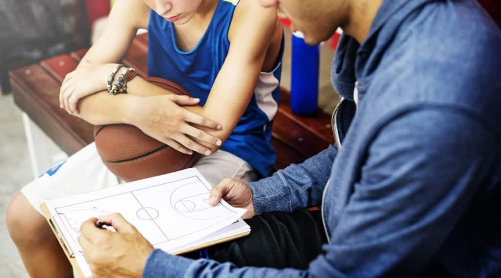 A sports coach sits with a basketball player, going over a play.