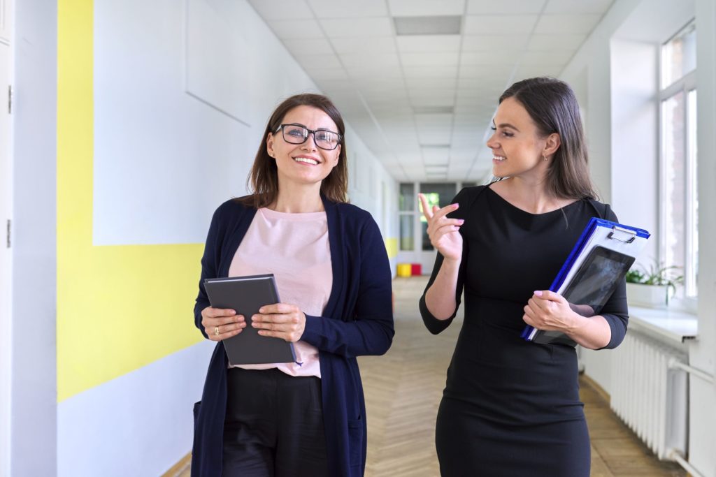 Two teachers walk and talk together down a hallway.