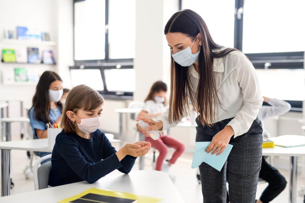 Masked teacher spraying sanitizer into the hands of a masked student.