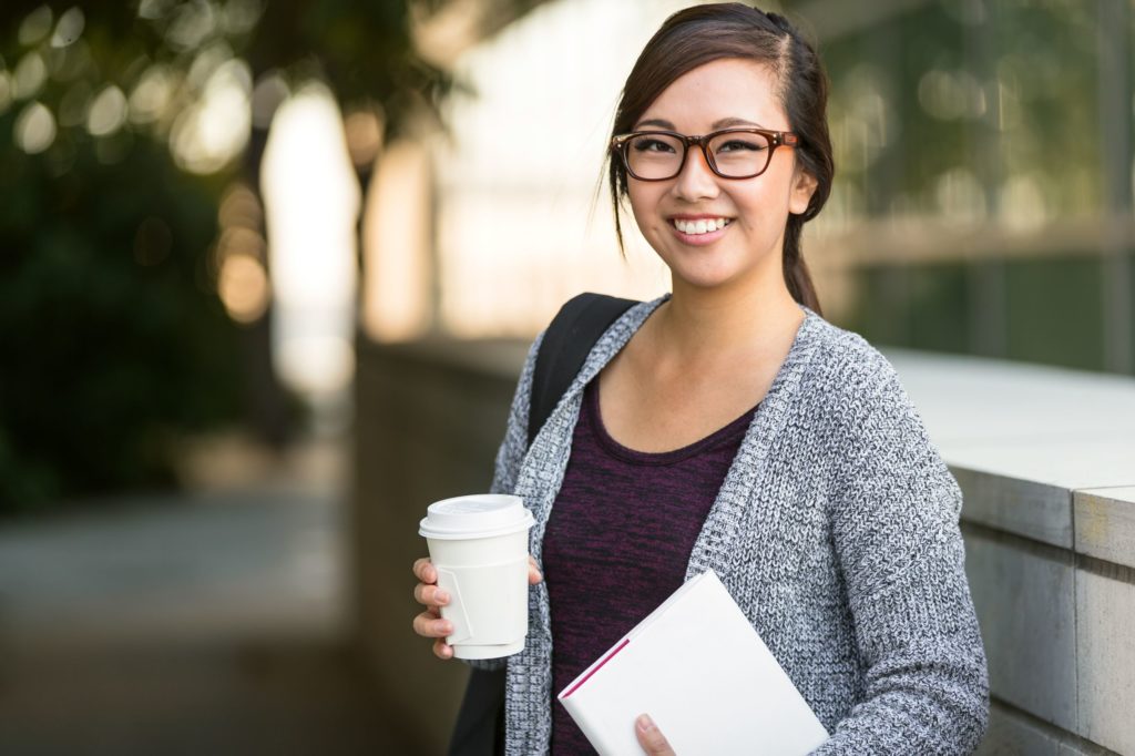 Student with backpack smiling and holding book and coffee.