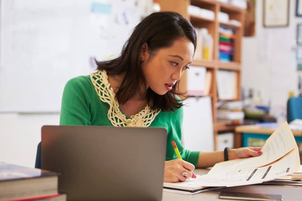 Young teacher taking notes while reviewing paperwork sitting at her desk.