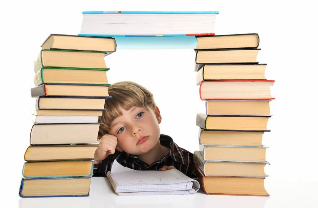 An upset school boy sits between towers of books.