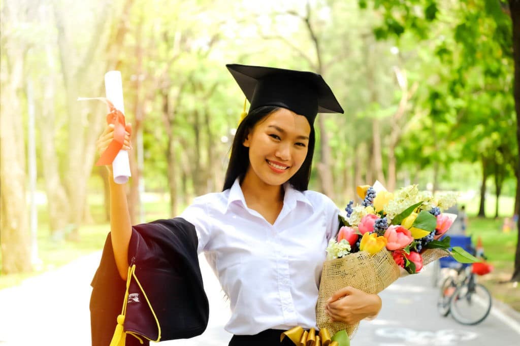A happy graduate in her cap stands holding flowers and her diploma.