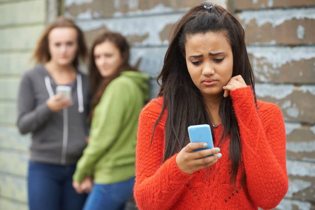 Sad girl looking at her cell phone with two girls staring behind her.