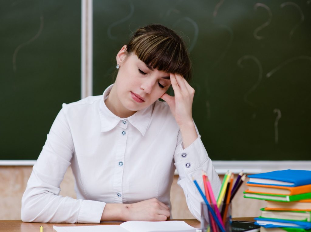 Stressed teacher with her hand on her head sitting at a desk.