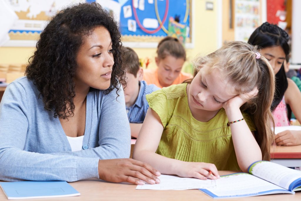 A teacher sits next to her struggling student as she assists her.