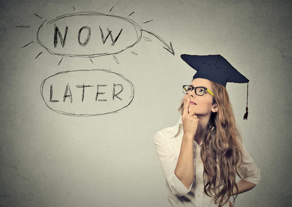 A woman wearing a graduation cap looks up at text that says “now” and “later”