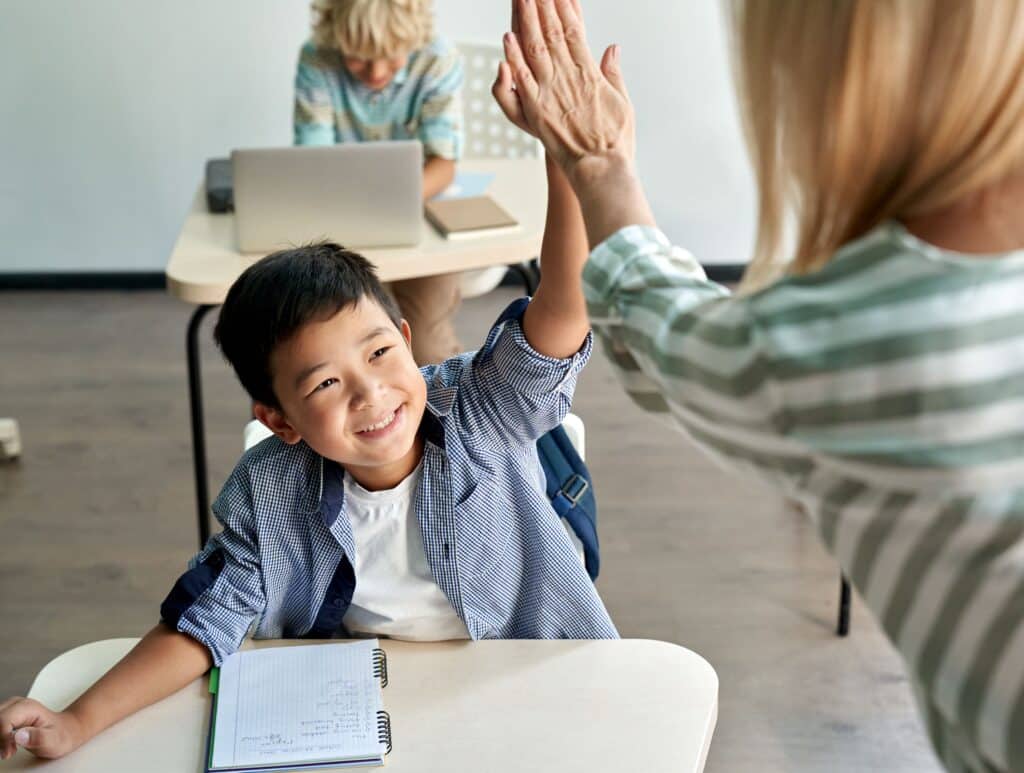 A young student gives a high-five to his teacher with a big smile because he has met his classroom goals.