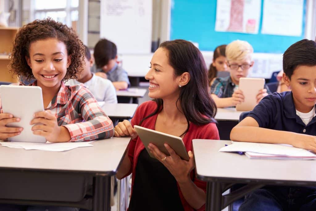 Teacher kneeling down next to a student at her desk and helping her.