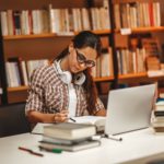 A woman sits in a library, studying with books and her laptop.