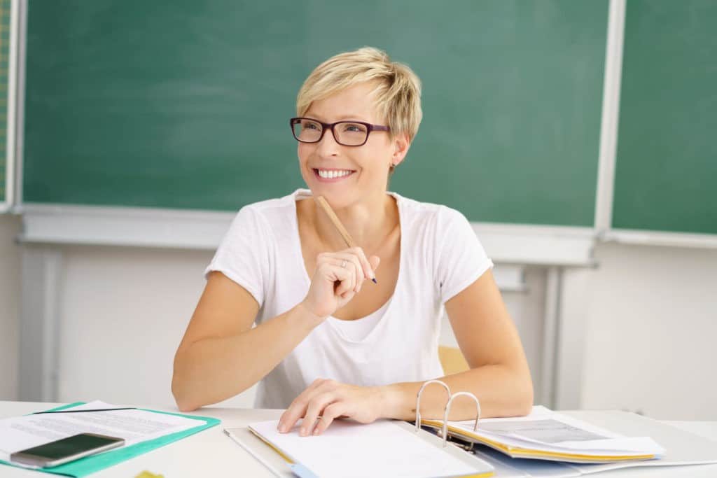 Smiling teacher sitting at a desk in front of a blackboard using a binder.