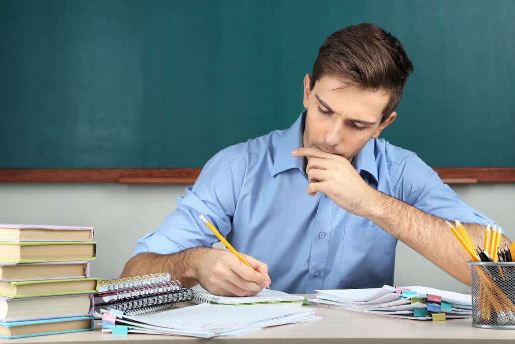 Teacher sitting at his desk in a classroom planning in a notebook.